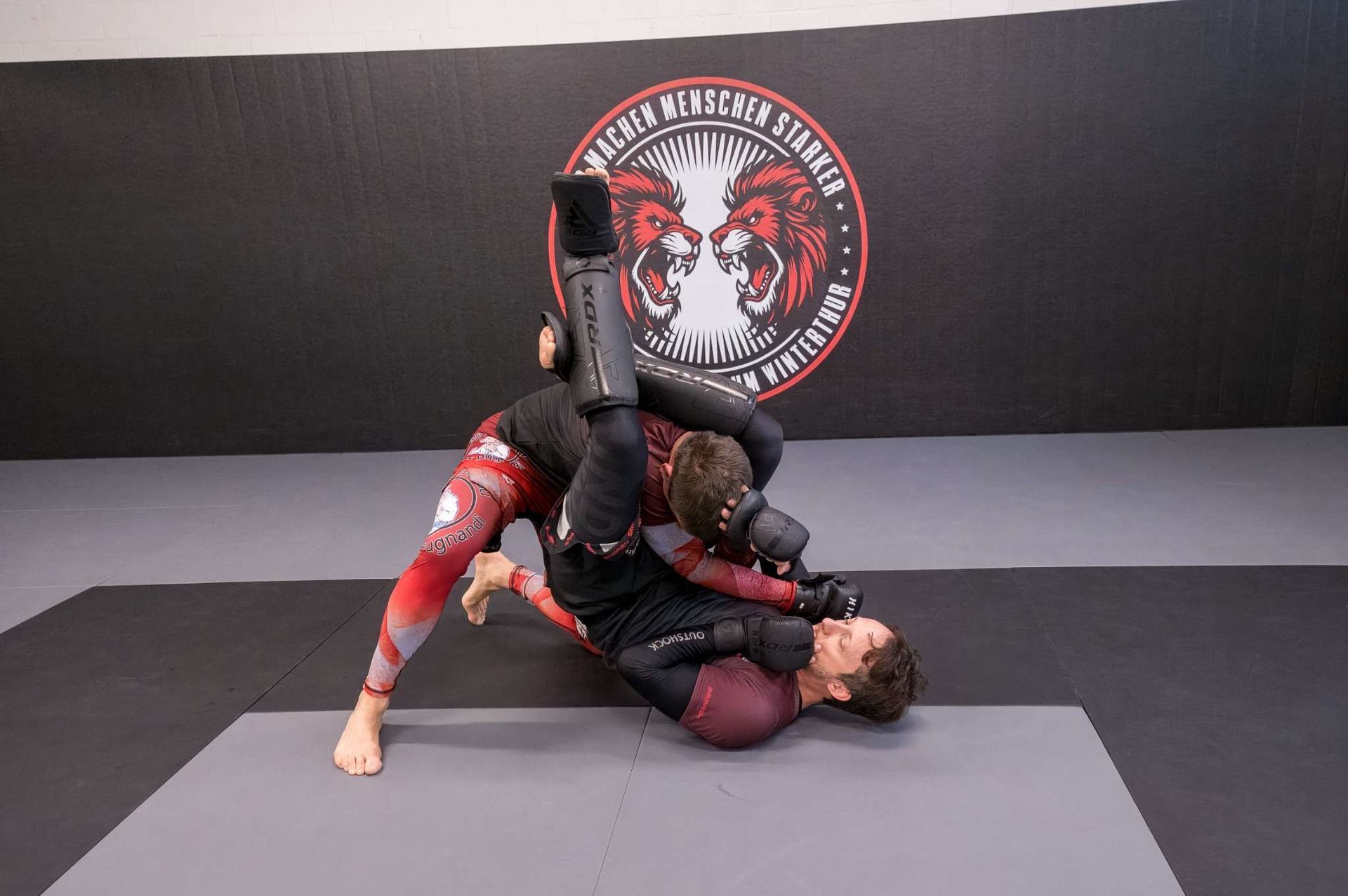 Ein Mann mit Bart bringt einem jungen Mann in einem Fitnessstudio Jiu-Jitsu bei.