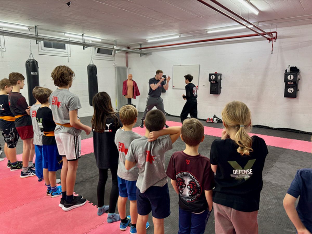 Eine Gruppe Kinder steht in einer Turnhalle und sieht einem Mann beim Boxen zu.