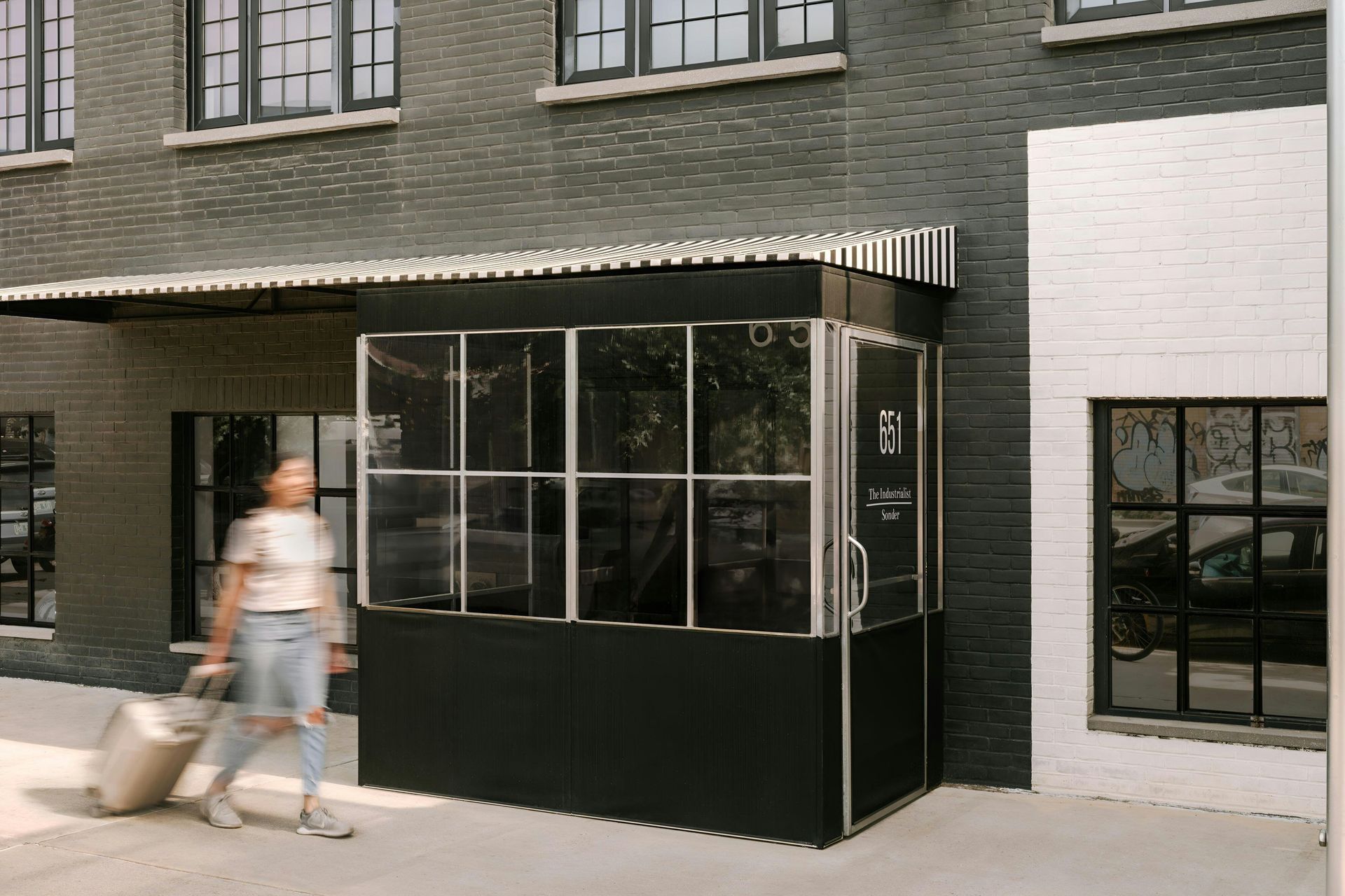 A person walks past a dark building with a modern metal and glass vestibule entrance, pulling a rolling suitcase.