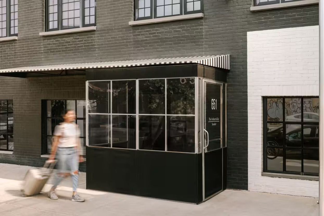 A person with a rolling suitcase walks past the black metal and glass entrance of a building with white-painted brick.