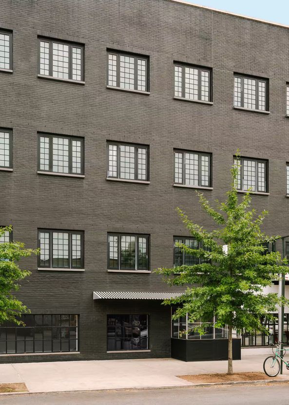 A multistory, dark-textured building exterior with a repeating wavy pattern, rectangular windows, and a street-level tree.