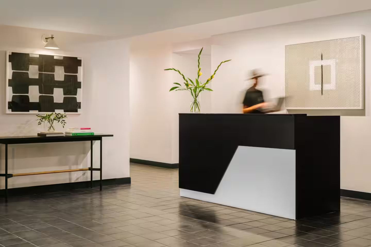 A modern hotel lobby with a black and white reception desk, minimalist art, and a blurred staff member at the counter.
