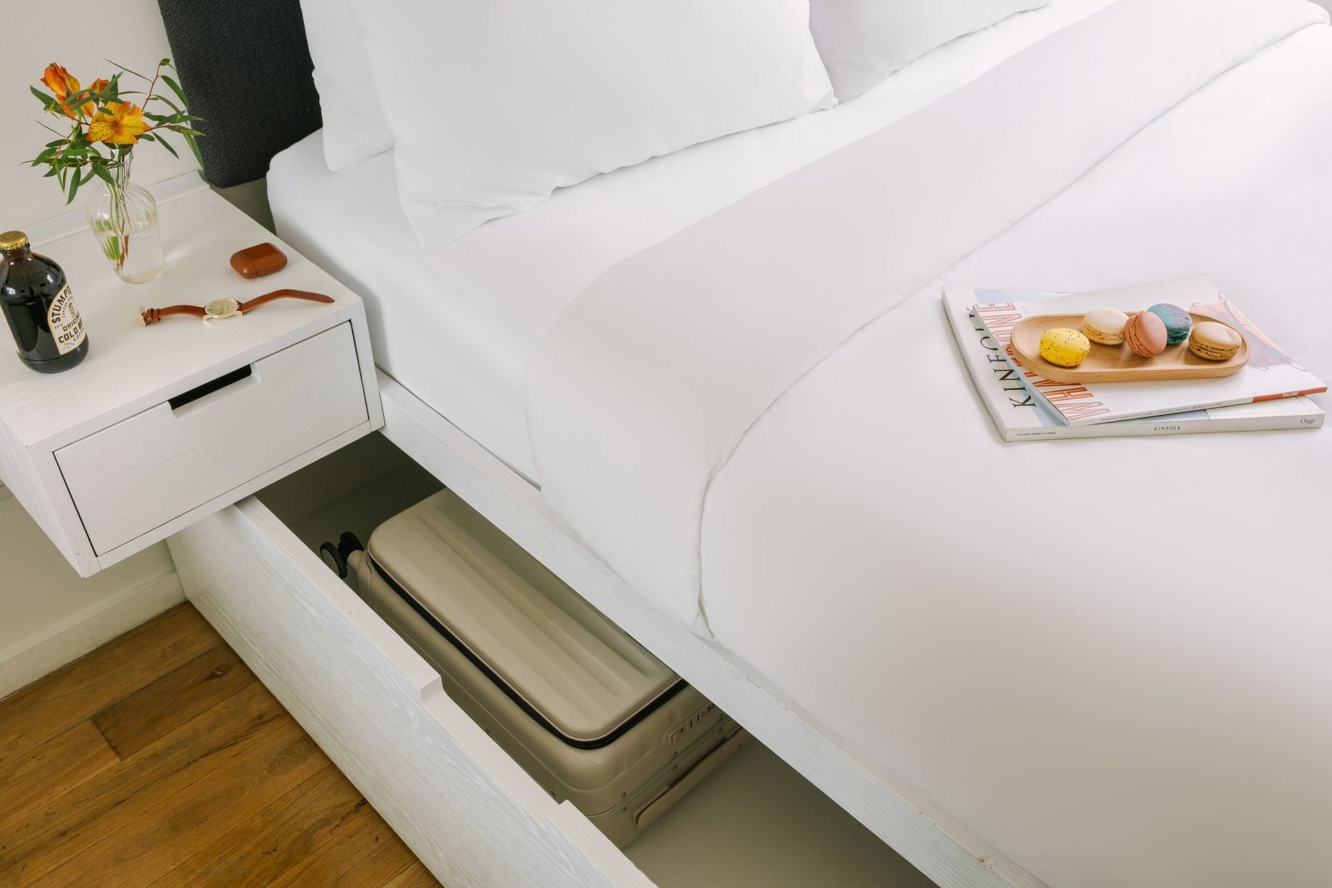 A white bed with an open under-bed drawer storing a suitcase, next to a matching white nightstand holding a small vase.