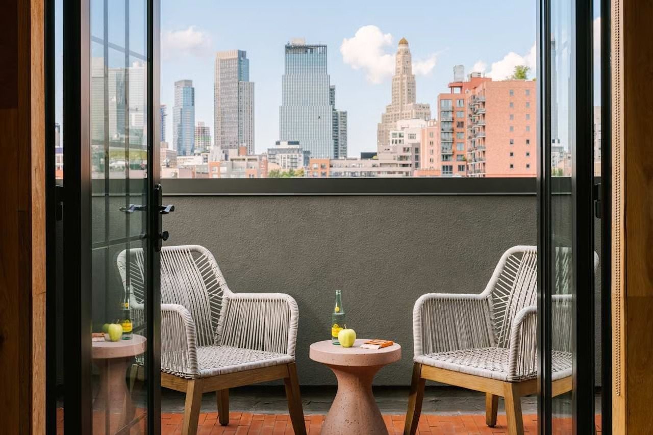 Two woven chairs and a small table with fruit on a balcony overlooking a city skyline on a sunny day.