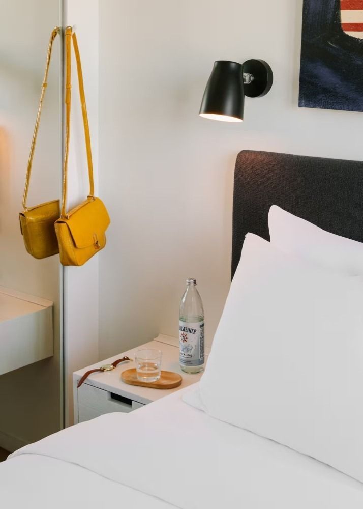 A bedroom interior featuring a white bed, a nightstand with a water bottle and glass, and a yellow bag hanging on a mirror.