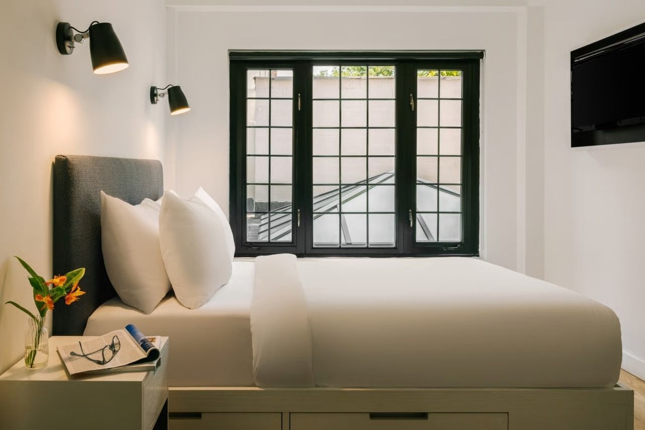 A white-walled hotel bedroom with a neatly made bed, black window frame, wall sconces, and a nightstand with flowers.