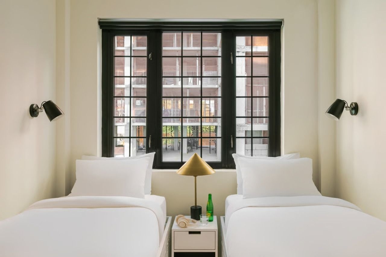 Two twin beds with white linens face a dark-framed window, centered by a small nightstand with a lamp and water bottle.