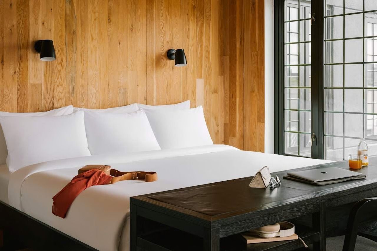 A bright bedroom with a white-made bed against a light wood-paneled wall, featuring two black wall lamps and a dark bench.