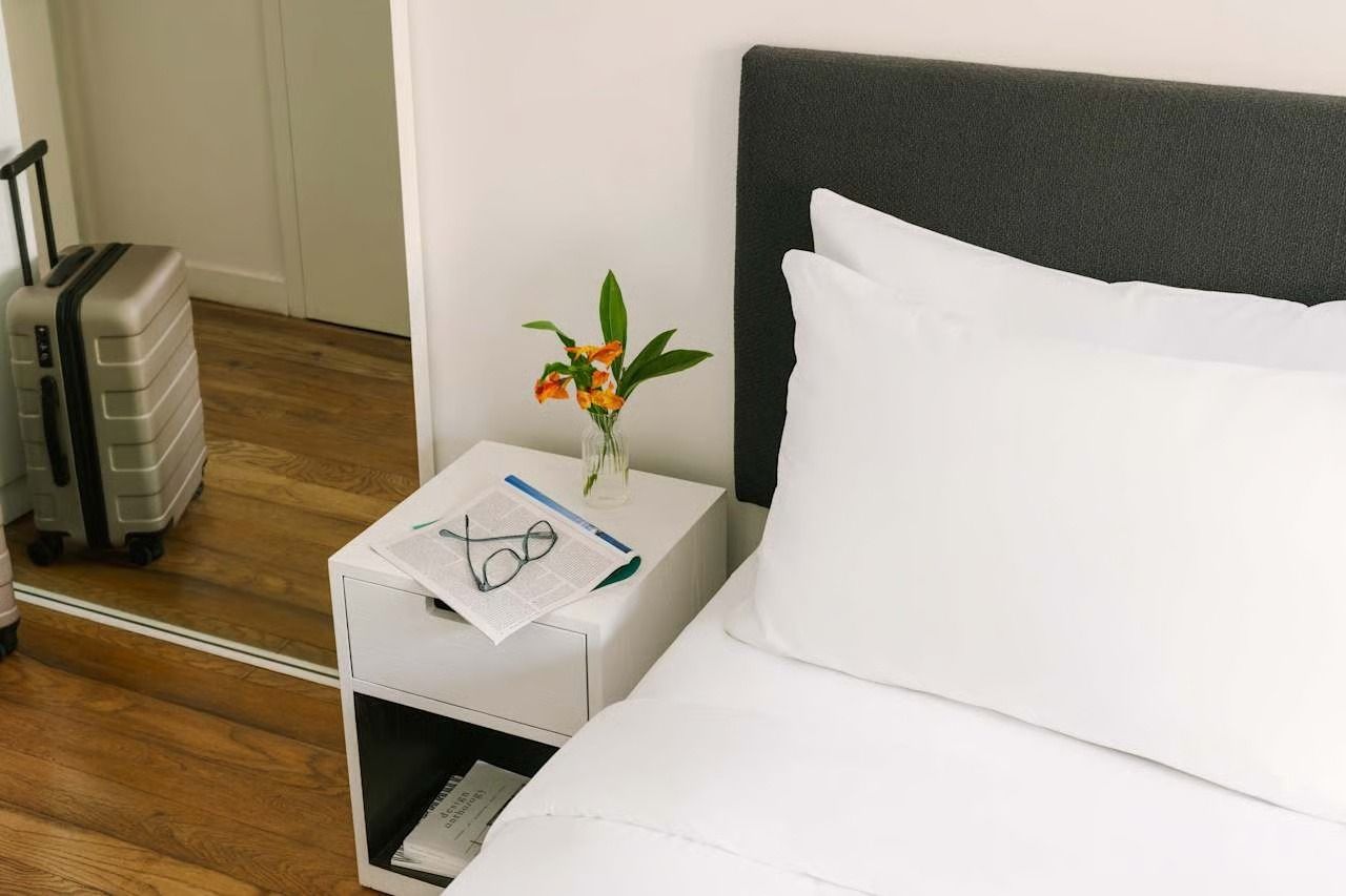 A bedside table with a small vase of flowers, glasses on a paper, and a grey suitcase nearby in a bedroom.