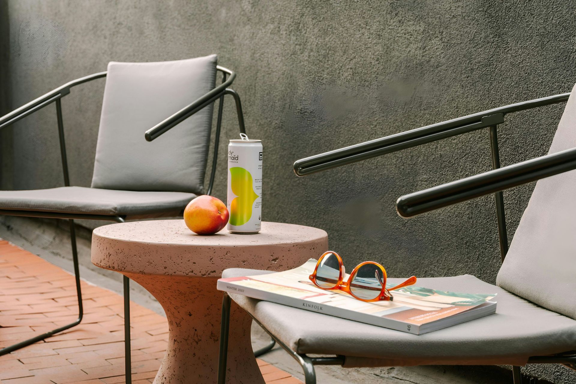 Two grey chairs and a stone table hold a drink can, an orange, and orange-rimmed sunglasses on a red brick patio.
