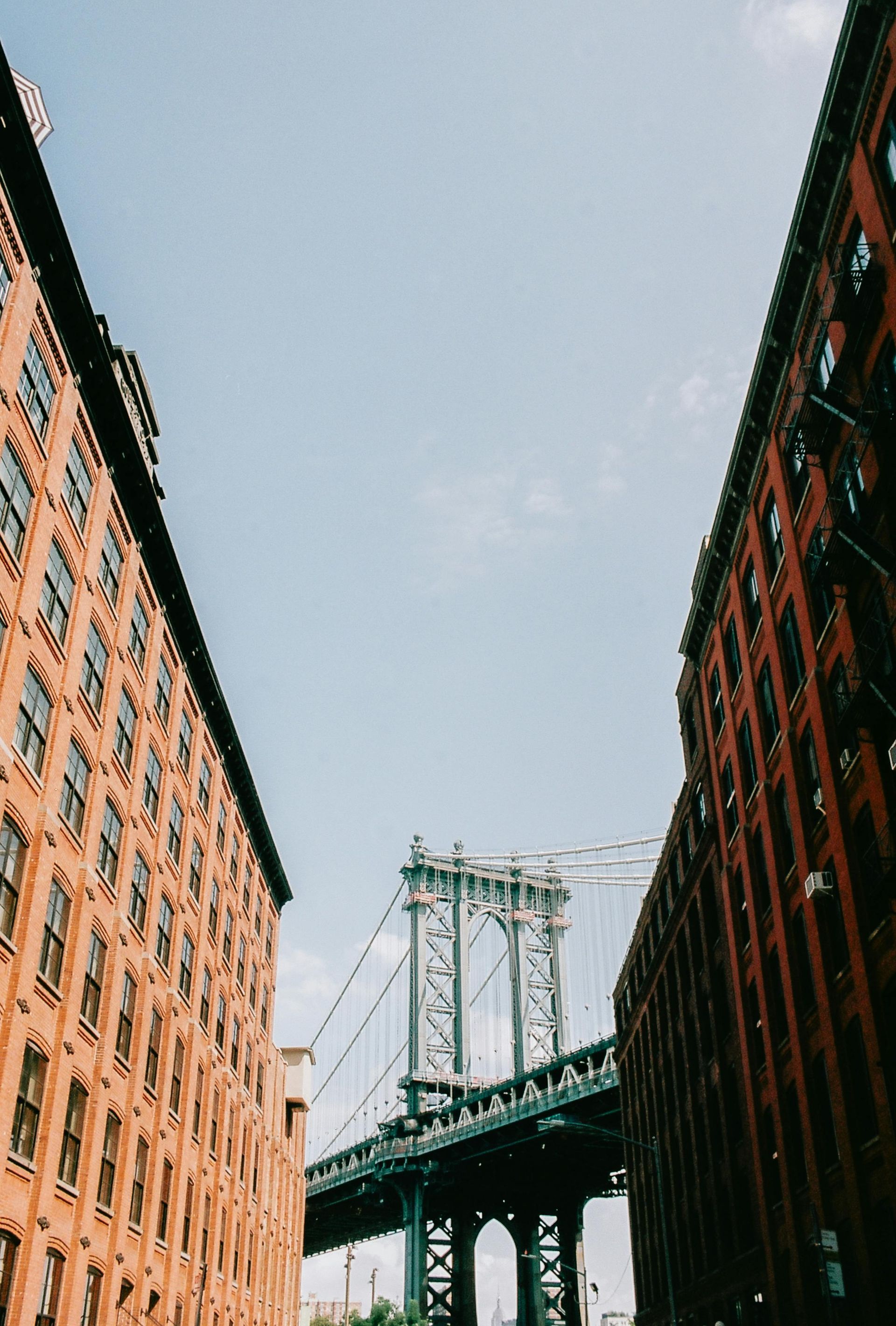 The Manhattan Bridge viewed from below between two tall, classic brick apartment buildings in New York City.