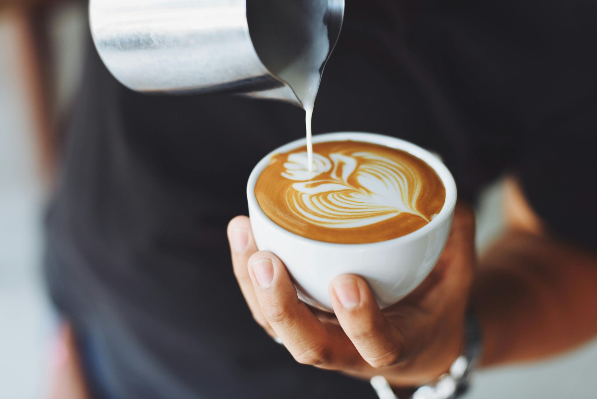 A person holding a white cup while pouring steamed milk into coffee, creating a delicate rosetta latte art design.