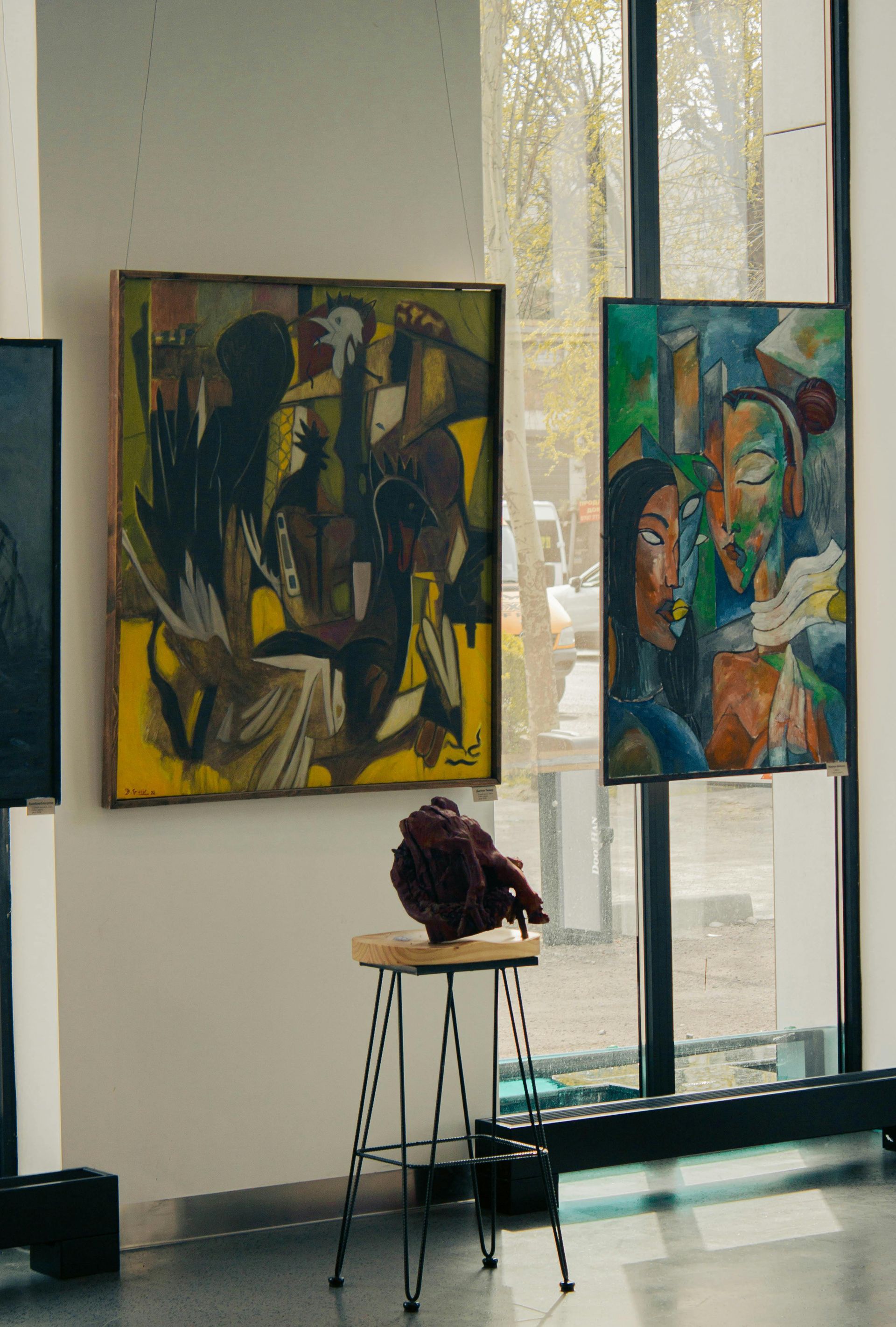 Two abstract paintings hang on a gallery wall near a window, with a small sculpture displayed on a stand in front.