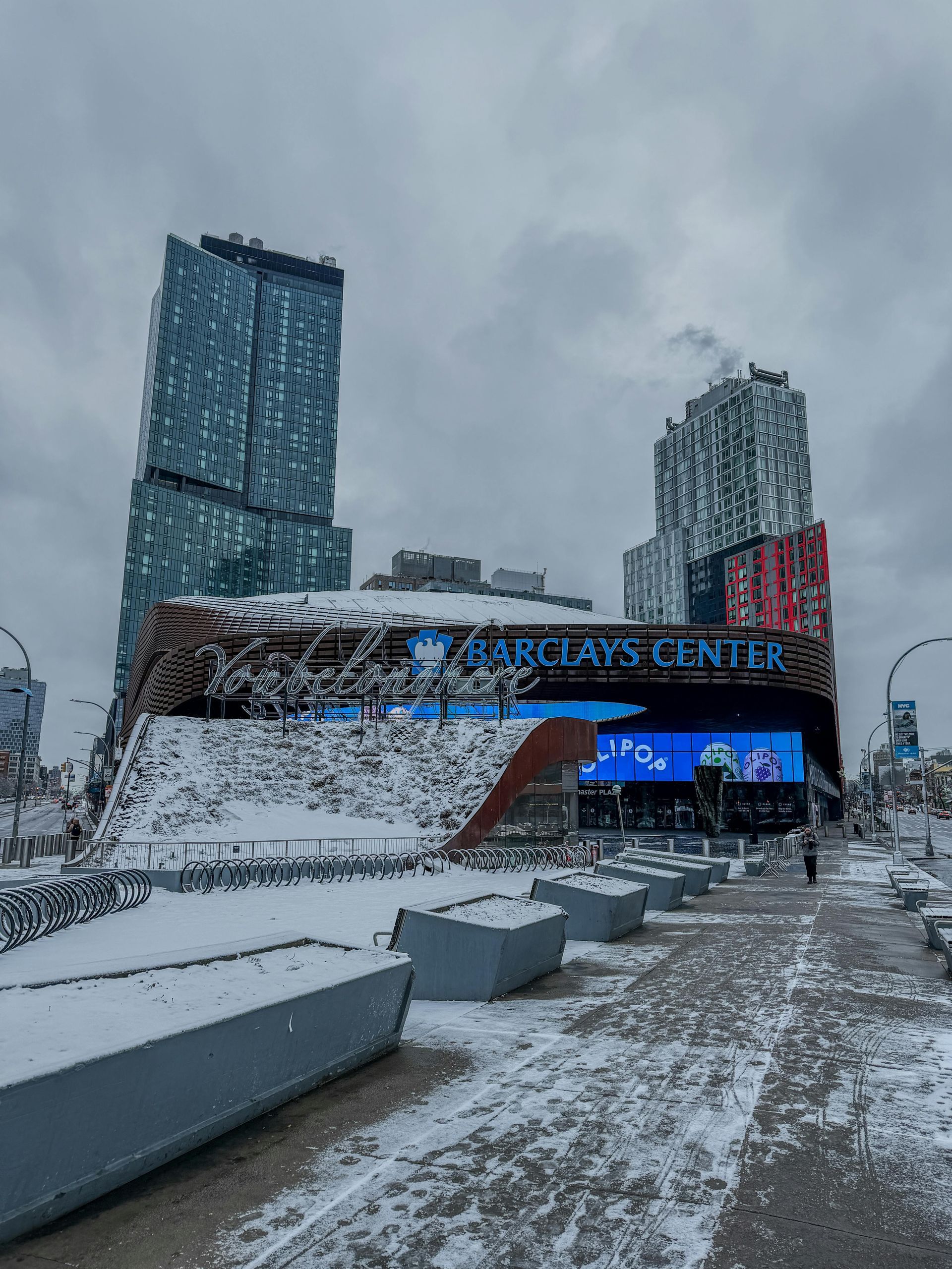 Snow covers the plaza in front of the Barclays Center in Brooklyn under a cloudy, overcast sky.