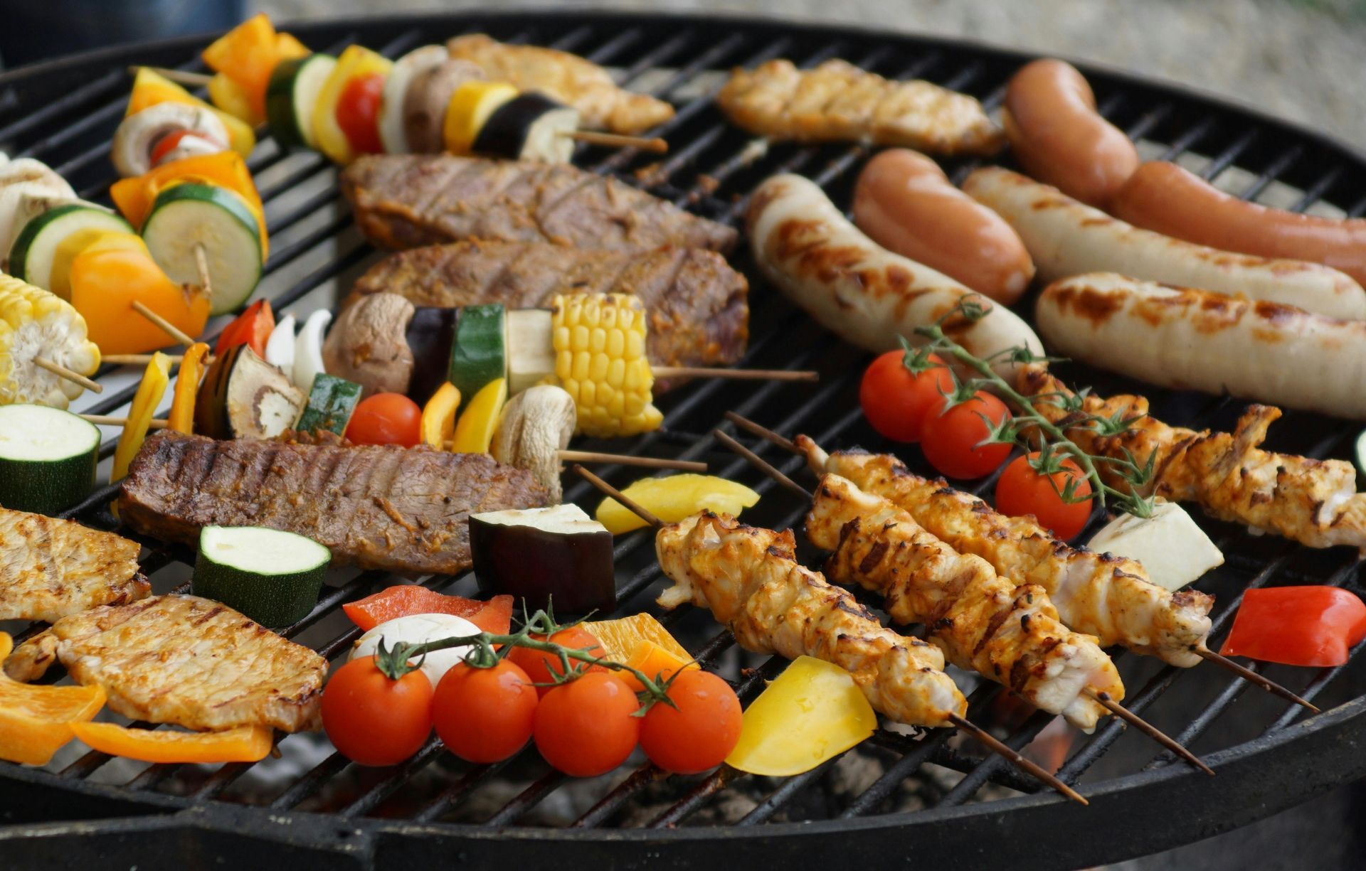 An assortment of skewered vegetables, meat, and sausages cooking on a black charcoal grill.