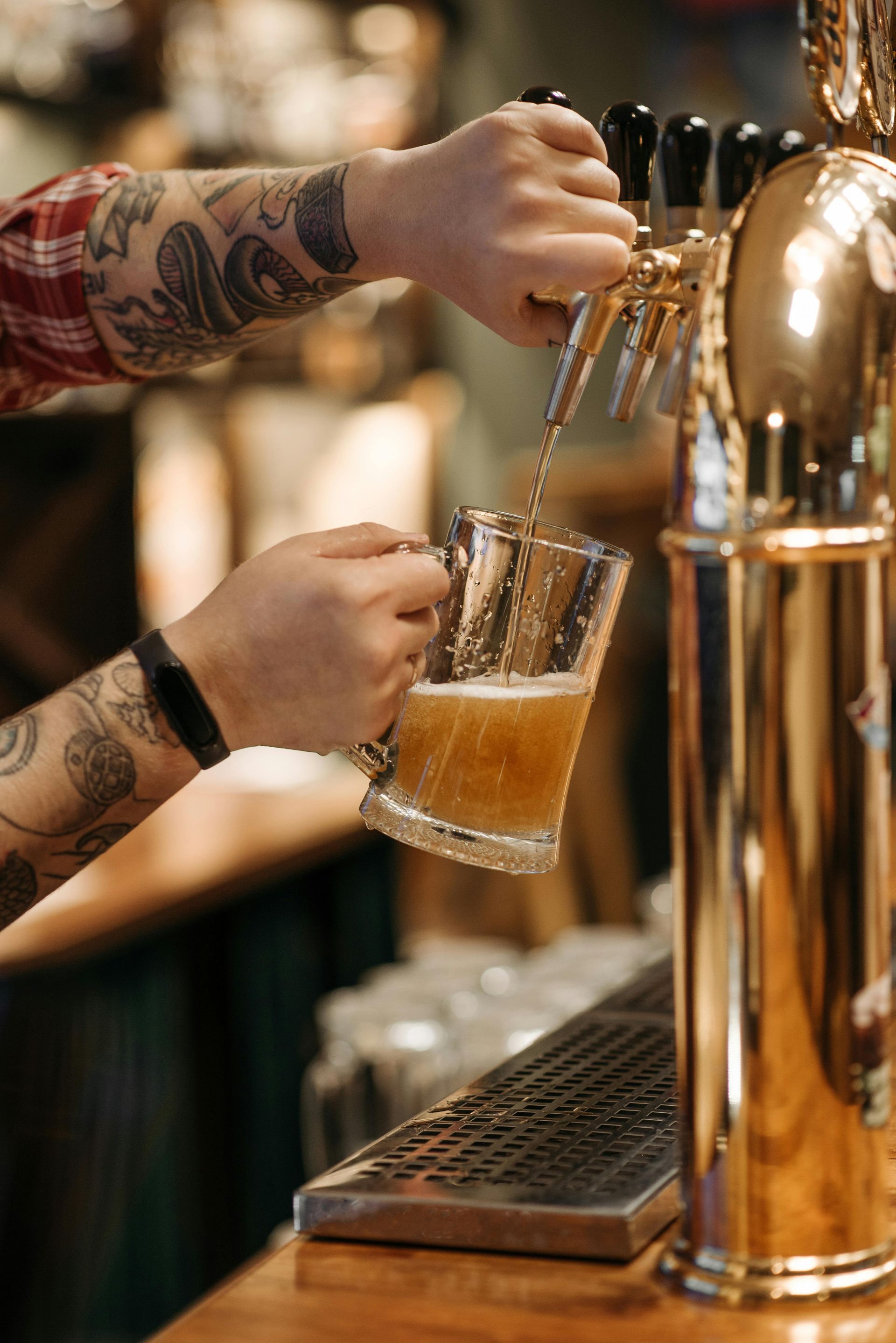 A tattooed person pours draft beer from a polished gold tap into a glass mug at a bar.