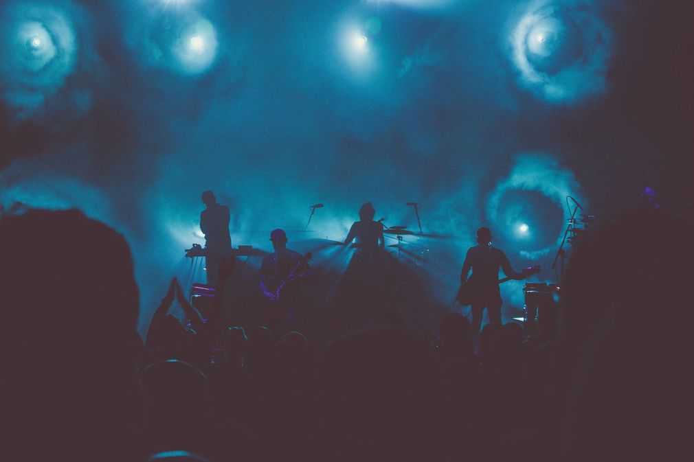 Silhouetted band members perform on a dimly lit stage filled with blue haze and bright, circular spotlights.