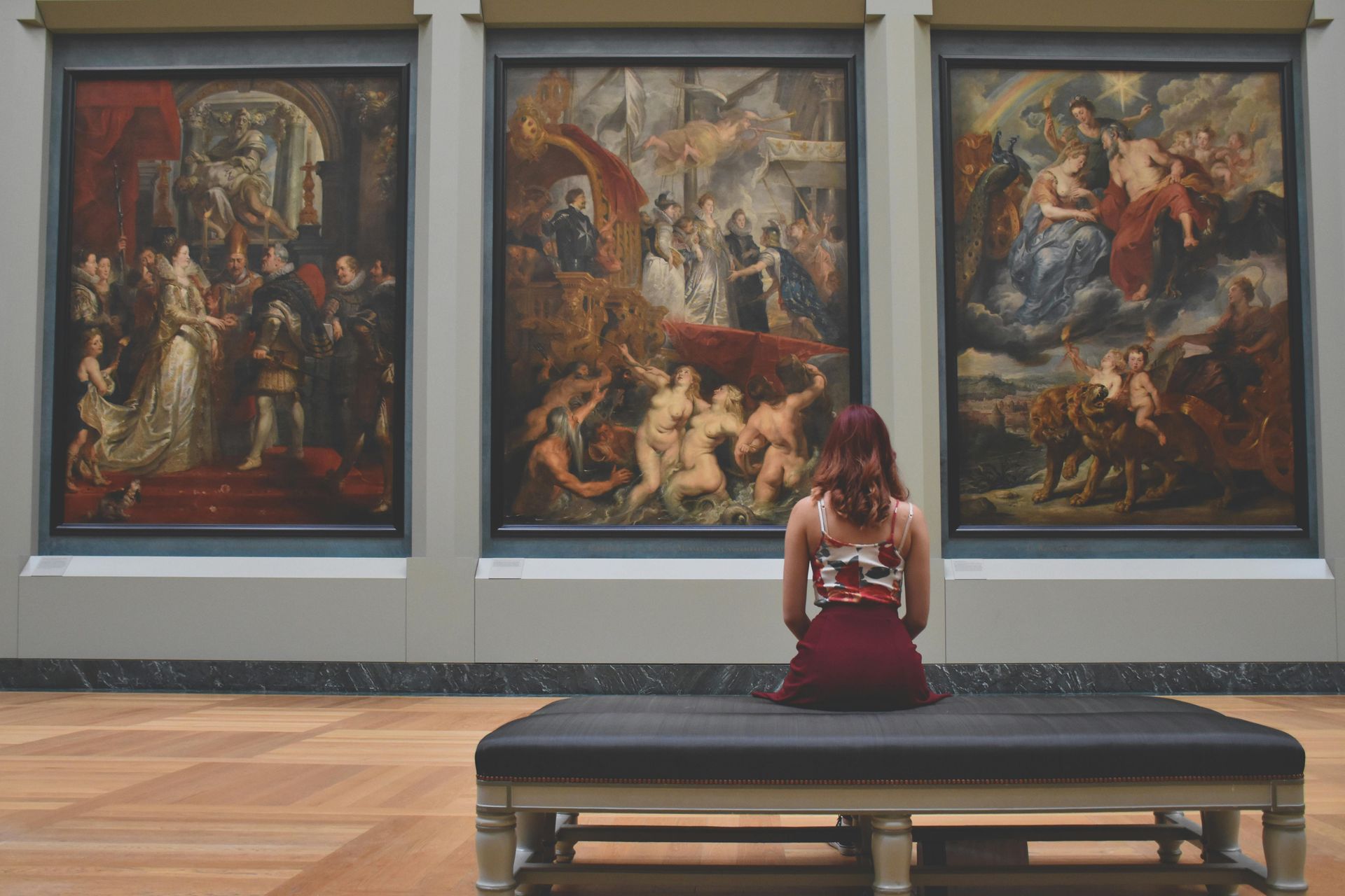 A person sits on a bench in an art gallery, facing three large, classical oil paintings displayed on a wall.