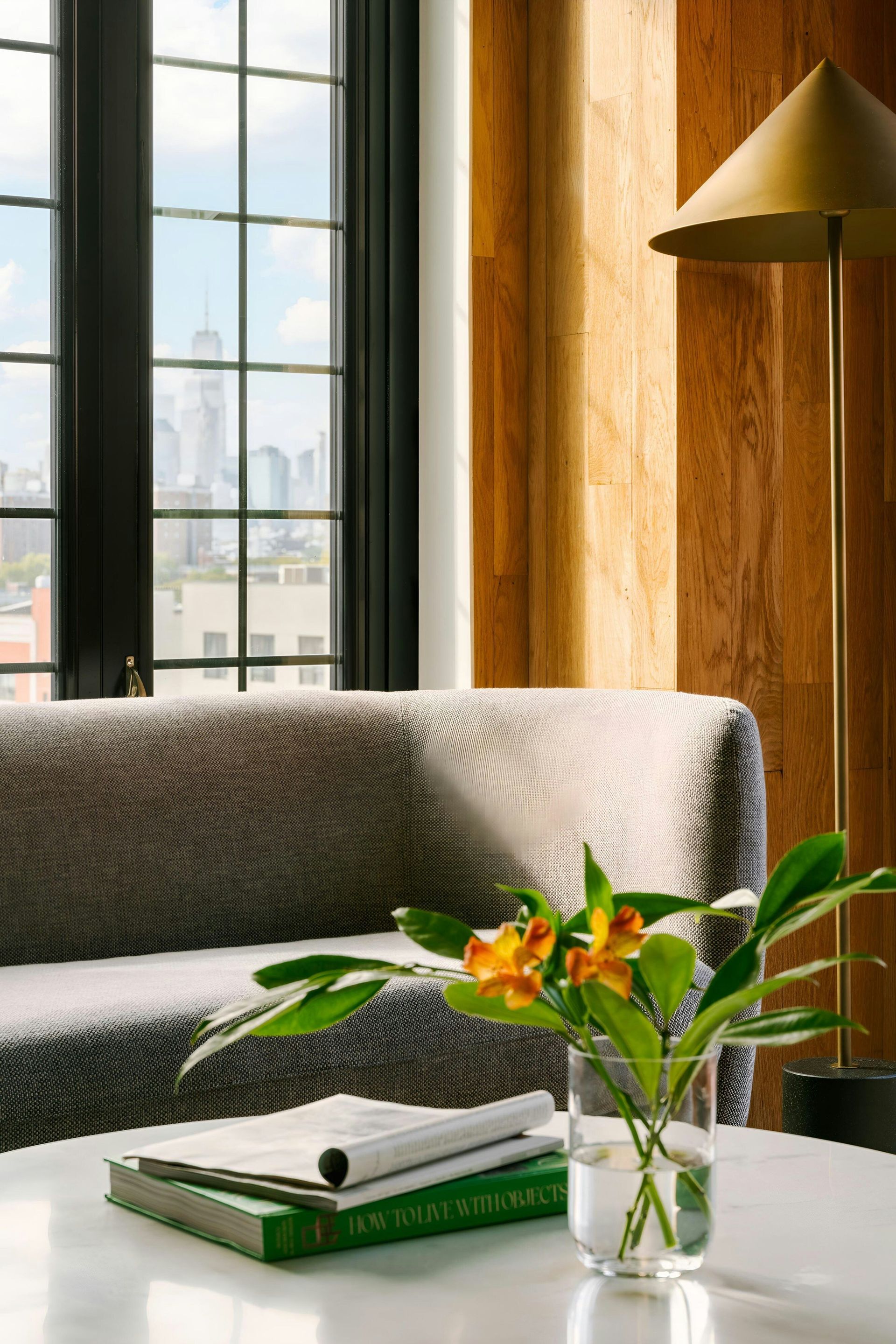 A patterned sofa, a vase with orange flowers, and books on a white table near a window with a city skyline view.