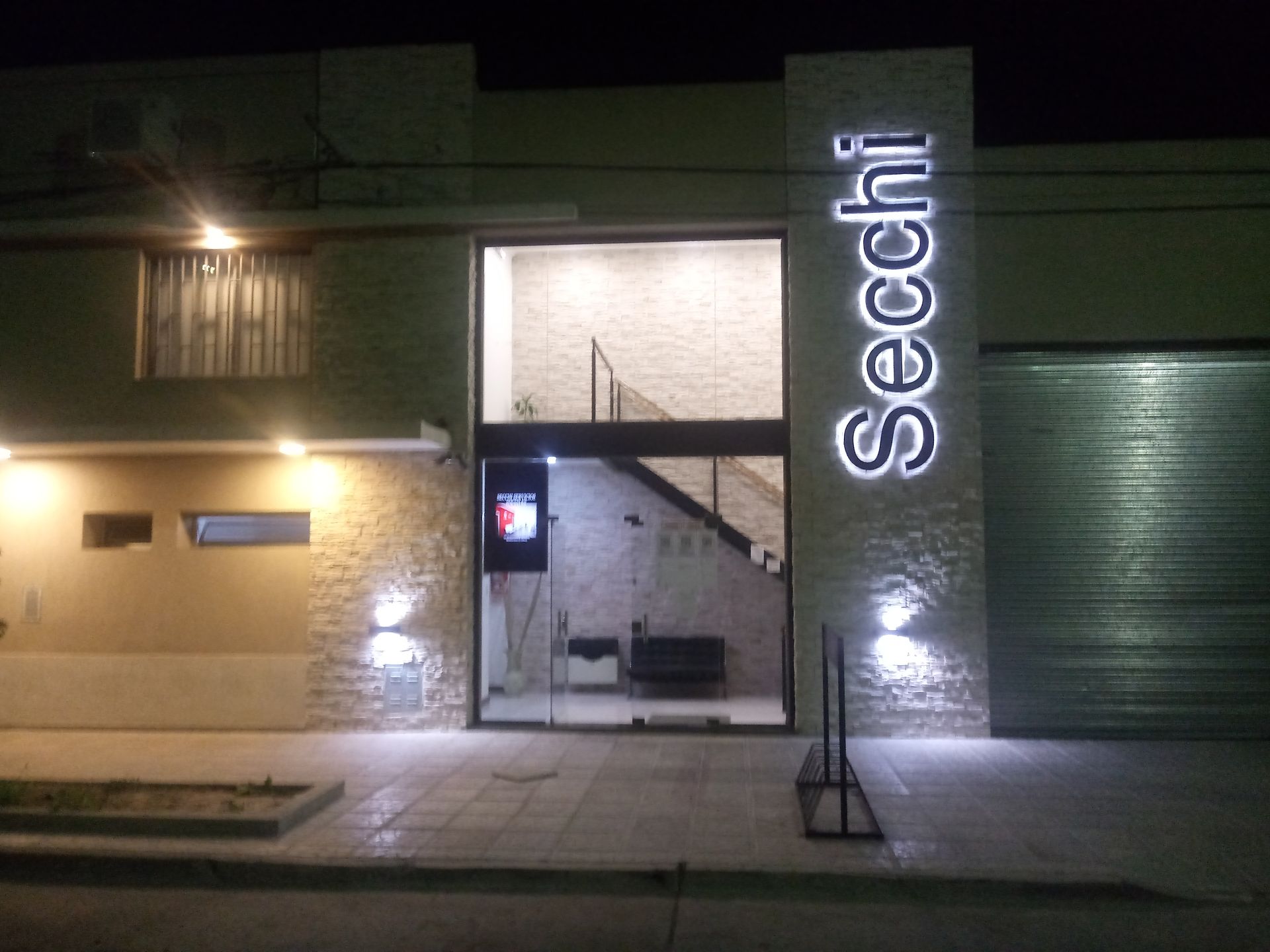 Por la noche, la fachada de un edificio presenta muros de piedra, una entrada central de cristal.