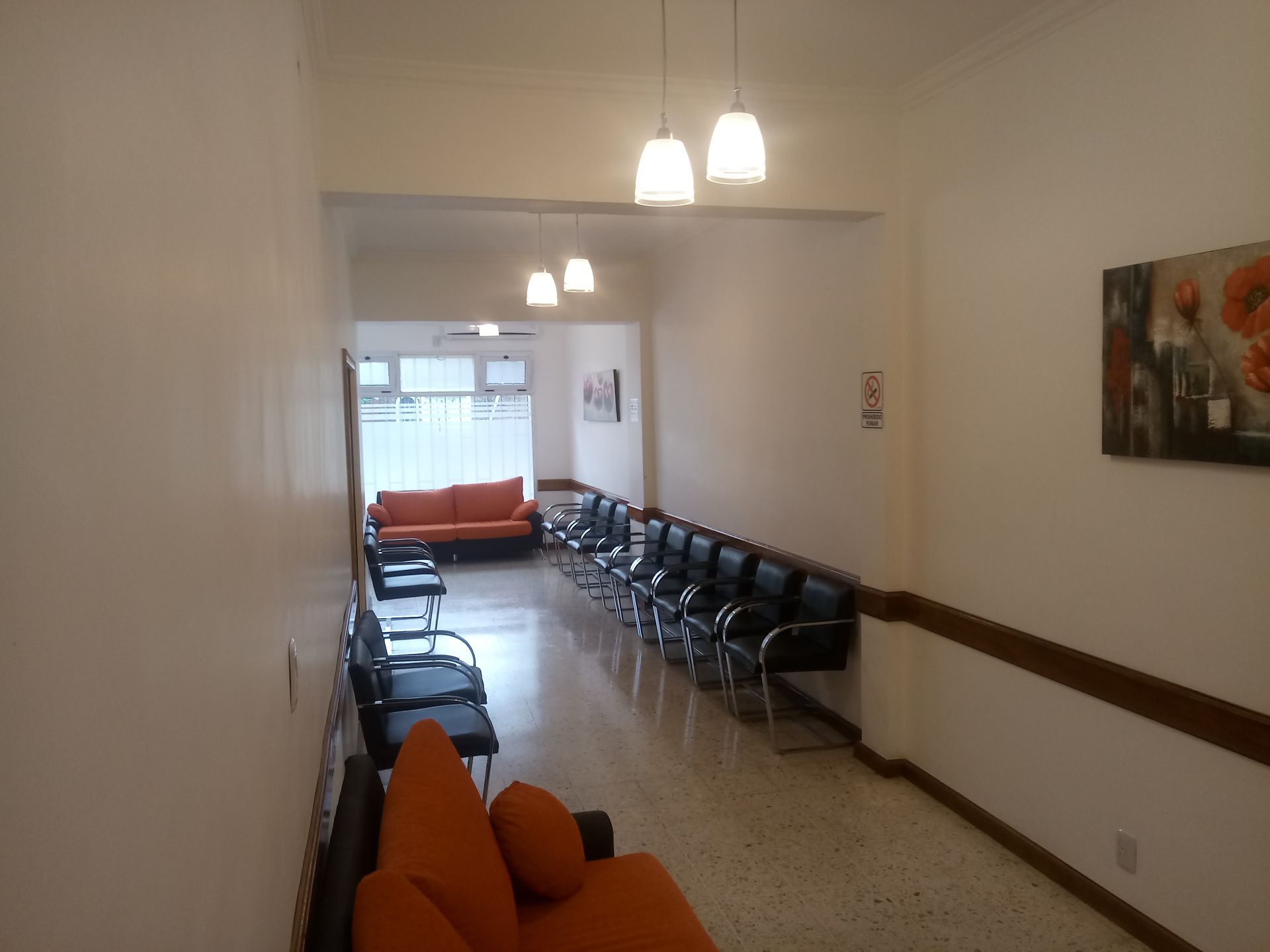 Una sala de espera con filas de sillas negras, dos sofás naranjas, suelo de baldosas y lámparas colgantes en un pasillo.
