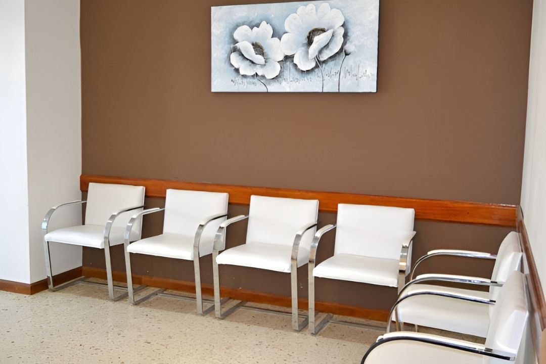 Una hilera de sillas blancas de sala de espera contra una pared marrón con un cuadro floral encima.