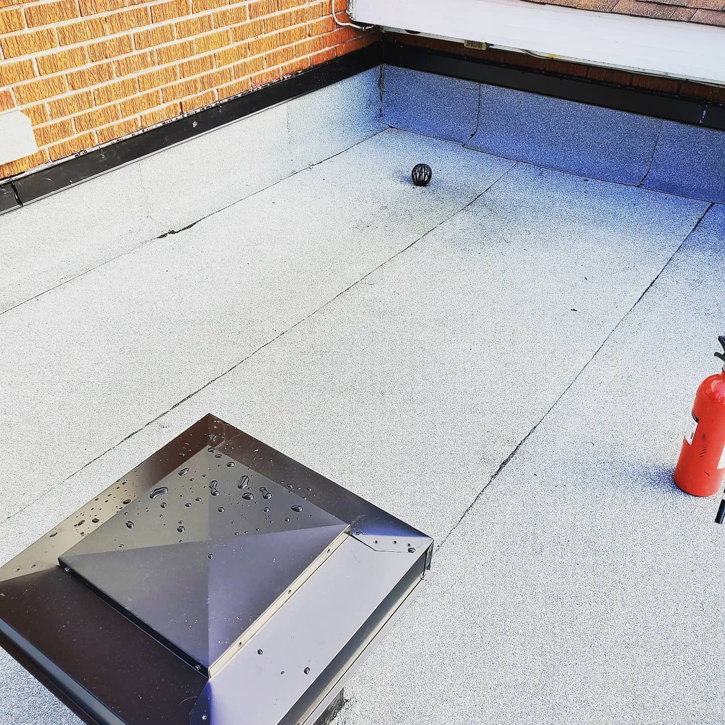 Toit plat à surface grise texturée. On distingue des garnitures noires, une cheminée marron et un extincteur rouge.