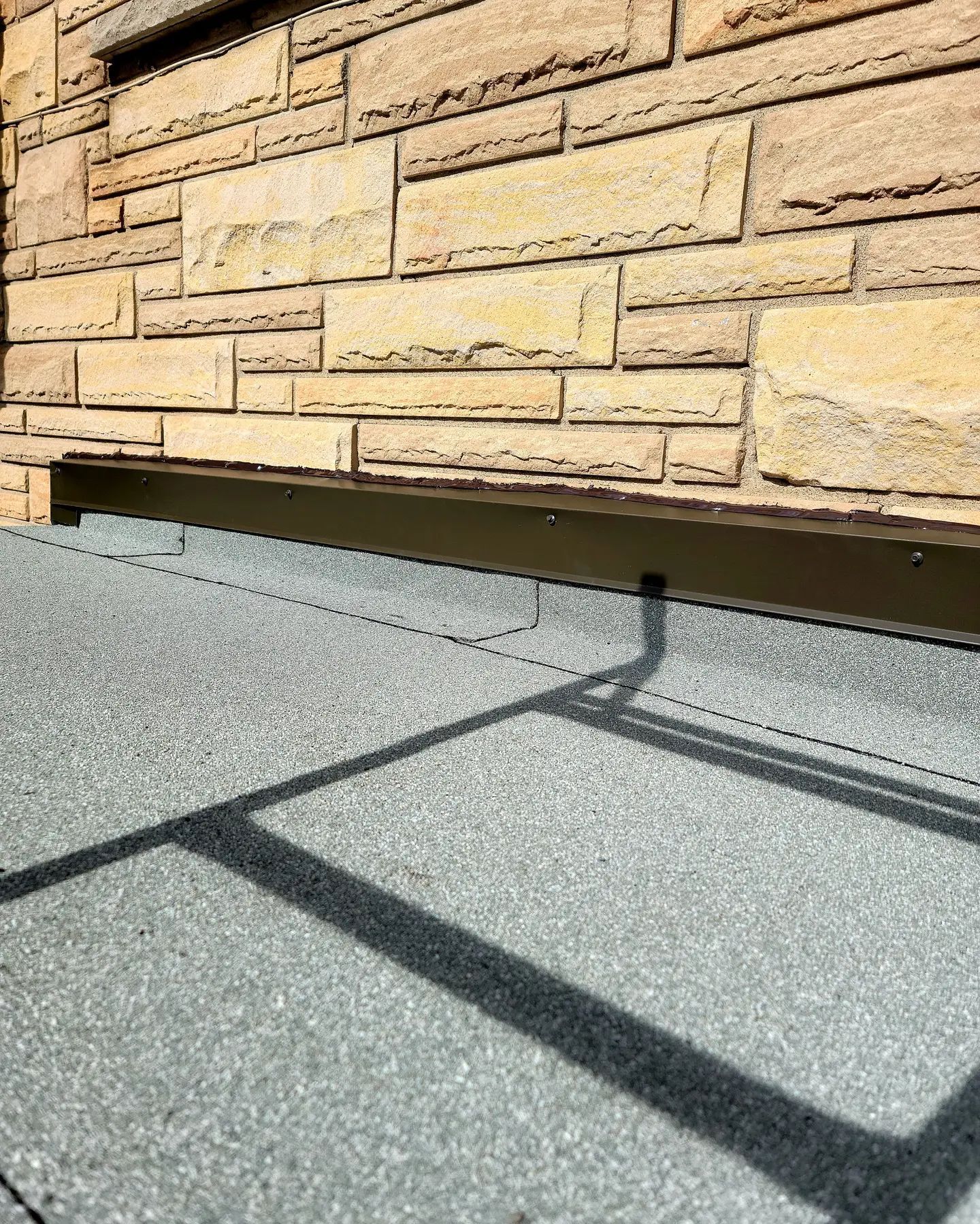 Mur de briques en pierre surplombant un toit plat et gris. Une barre métallique sombre s'étend du mur.