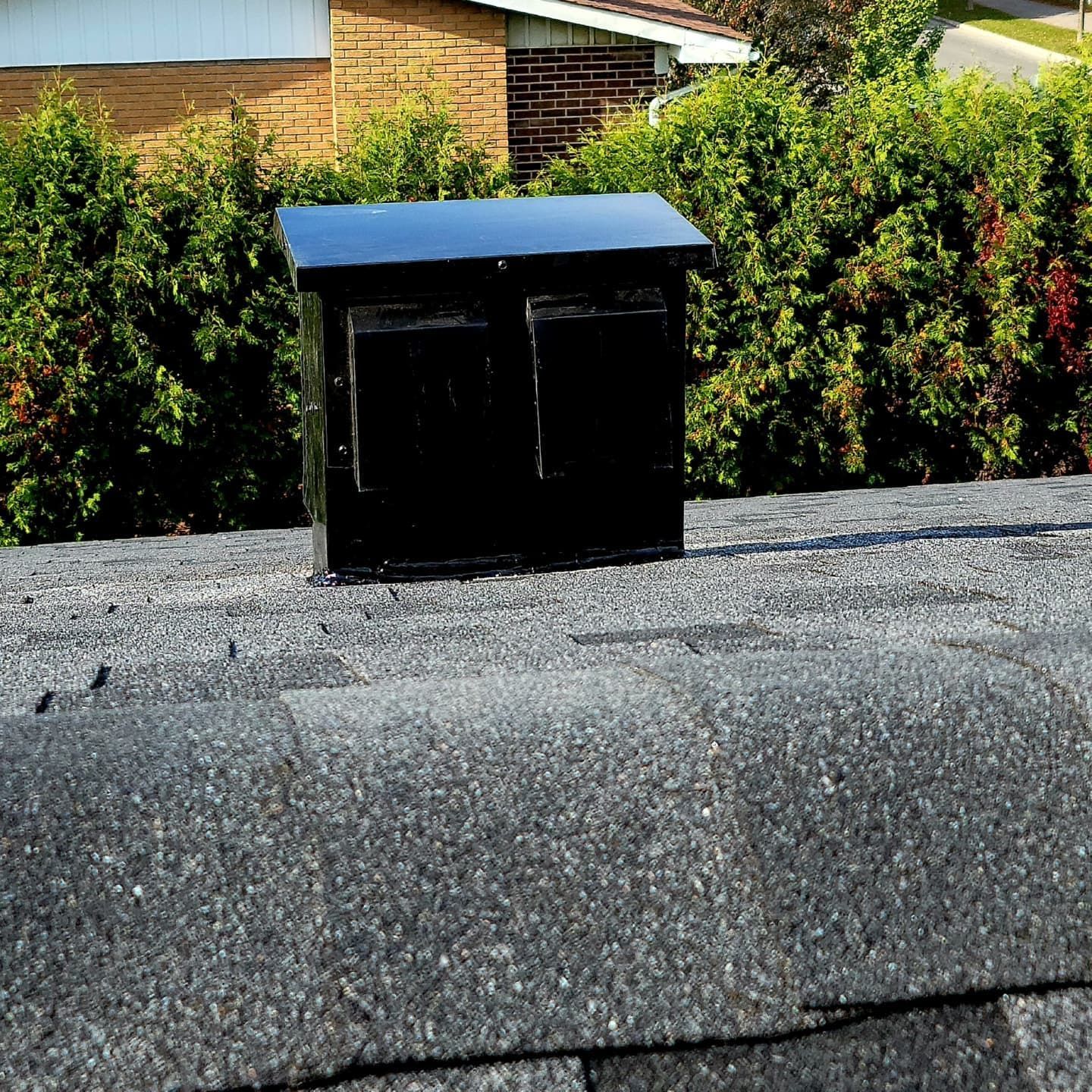 Évent de toit noir sur un toit en bardeaux avec un arrière-plan de buissons verts.