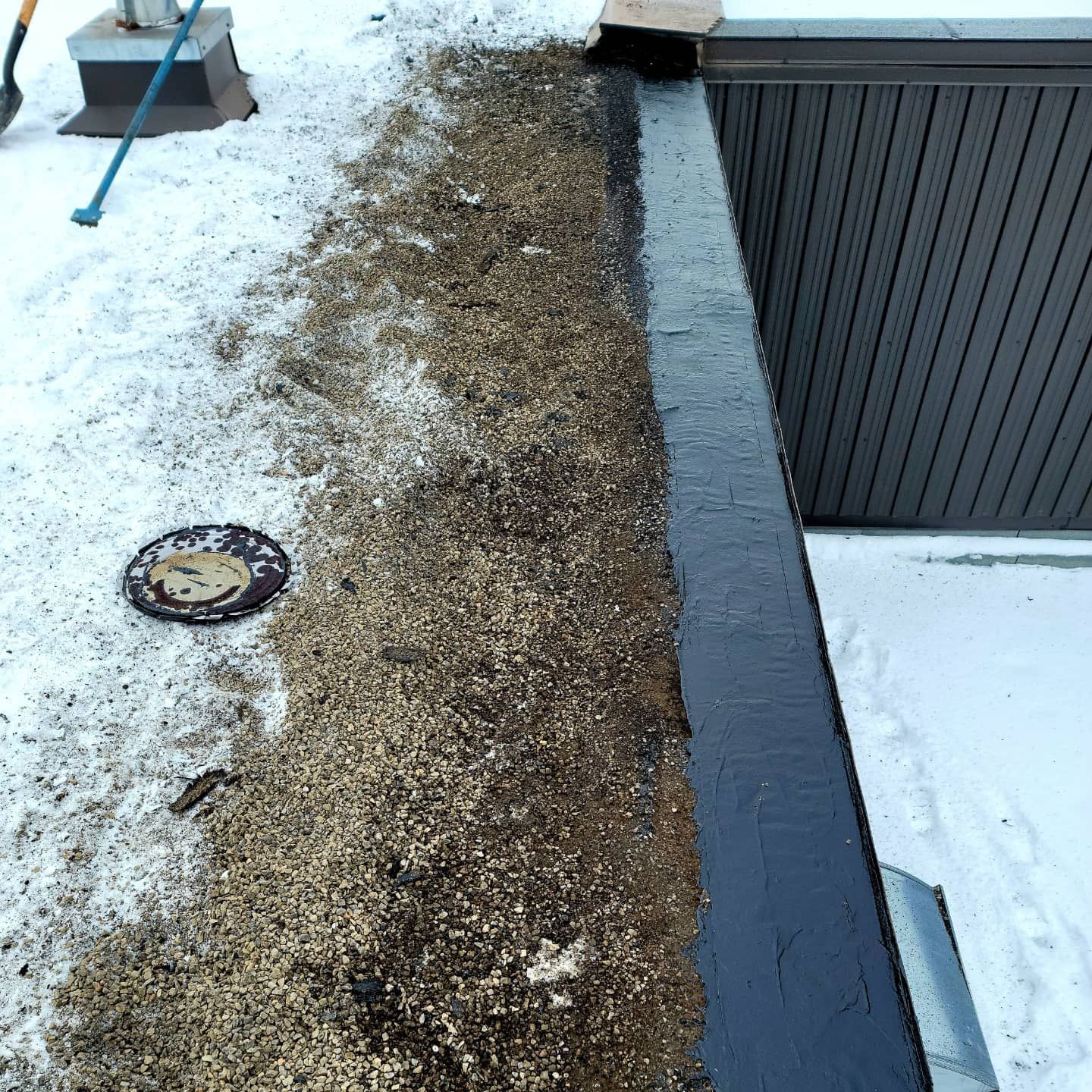 Toit plat recouvert de gravier, de neige et de mastic noir. On aperçoit le bord d'un bâtiment.