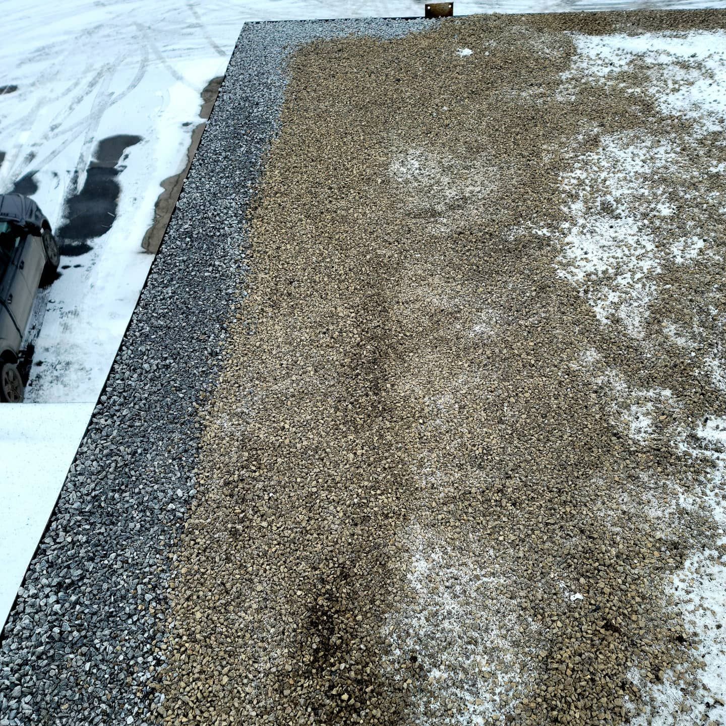 Toit plat avec gravier gris et beige, un peu de neige et bord près d'une voiture.