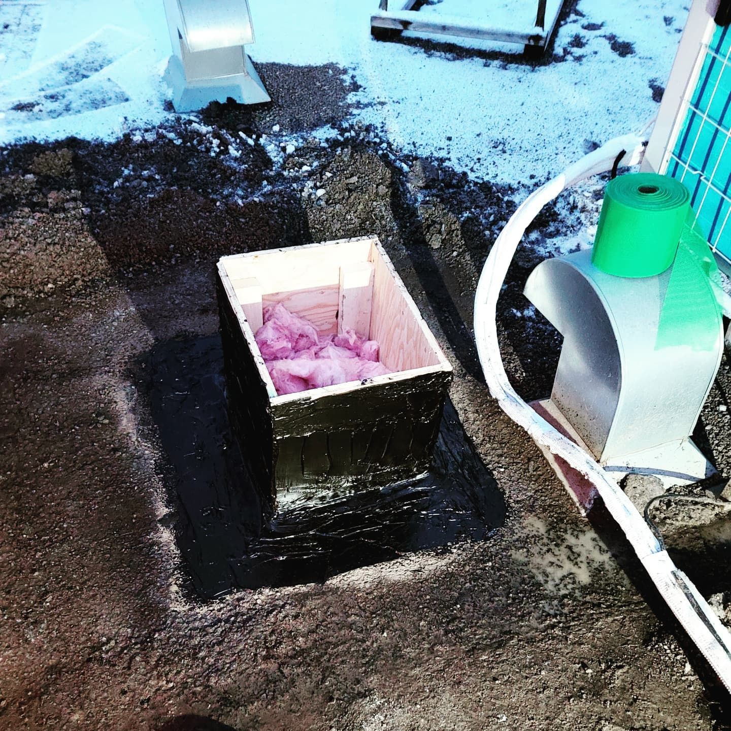 Boîte ouverte avec isolation rose sur un toit en goudron, à côté de ruban vert et d'un panneau solaire.