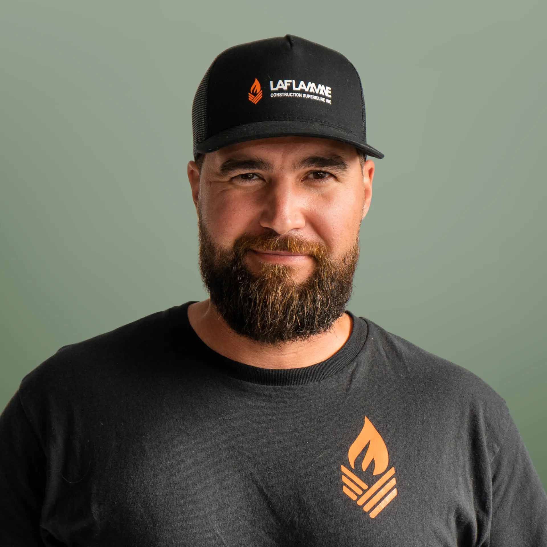 Un homme barbu portant une casquette noire et un t-shirt avec un logo de flamme sourit à la caméra.