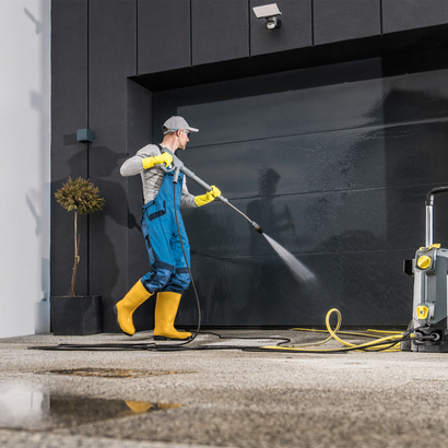 Man pressure washing a dark garage door wearing yellow boots, gloves, and a blue jumpsuit.