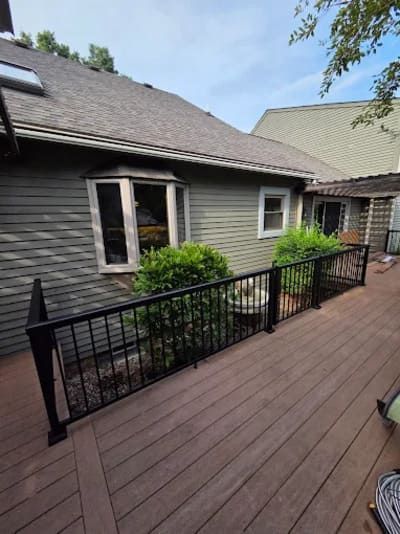 A wooden deck with a black metal railing, adjacent to a gray house with a bay window and green bushes.