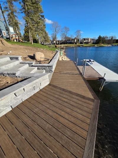 Wooden deck leading to a metal dock on a lake with tiered stone steps and a retaining wall on the grassy shoreline.