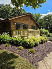 A rustic log cabin with a wooden deck surrounded by green shrubs and dark mulch under a bright blue sky.