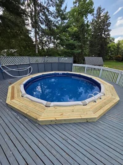 A round above-ground pool surrounded by a custom octagonal wood deck on a gray wooden patio in a backyard.