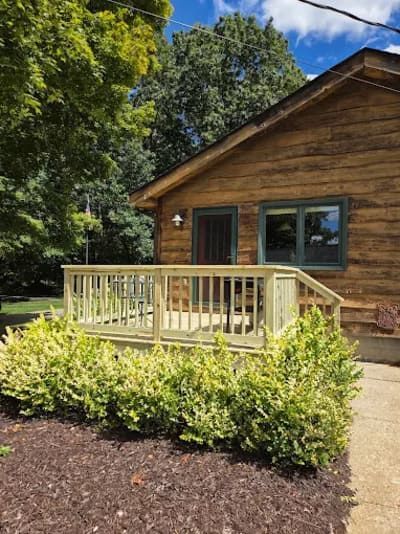 A rustic log cabin with a light-colored wooden deck, framed by green landscaping and mulch, set against a sunny sky.