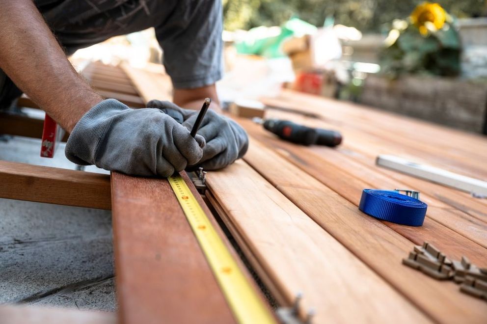 Hands wearing gray work gloves use a pencil and tape measure to mark wood planks on a deck construction site.