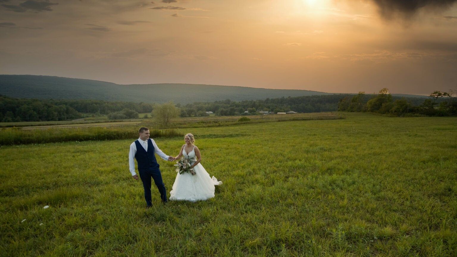 A bride and groom are walking through a grassy field at sunset.