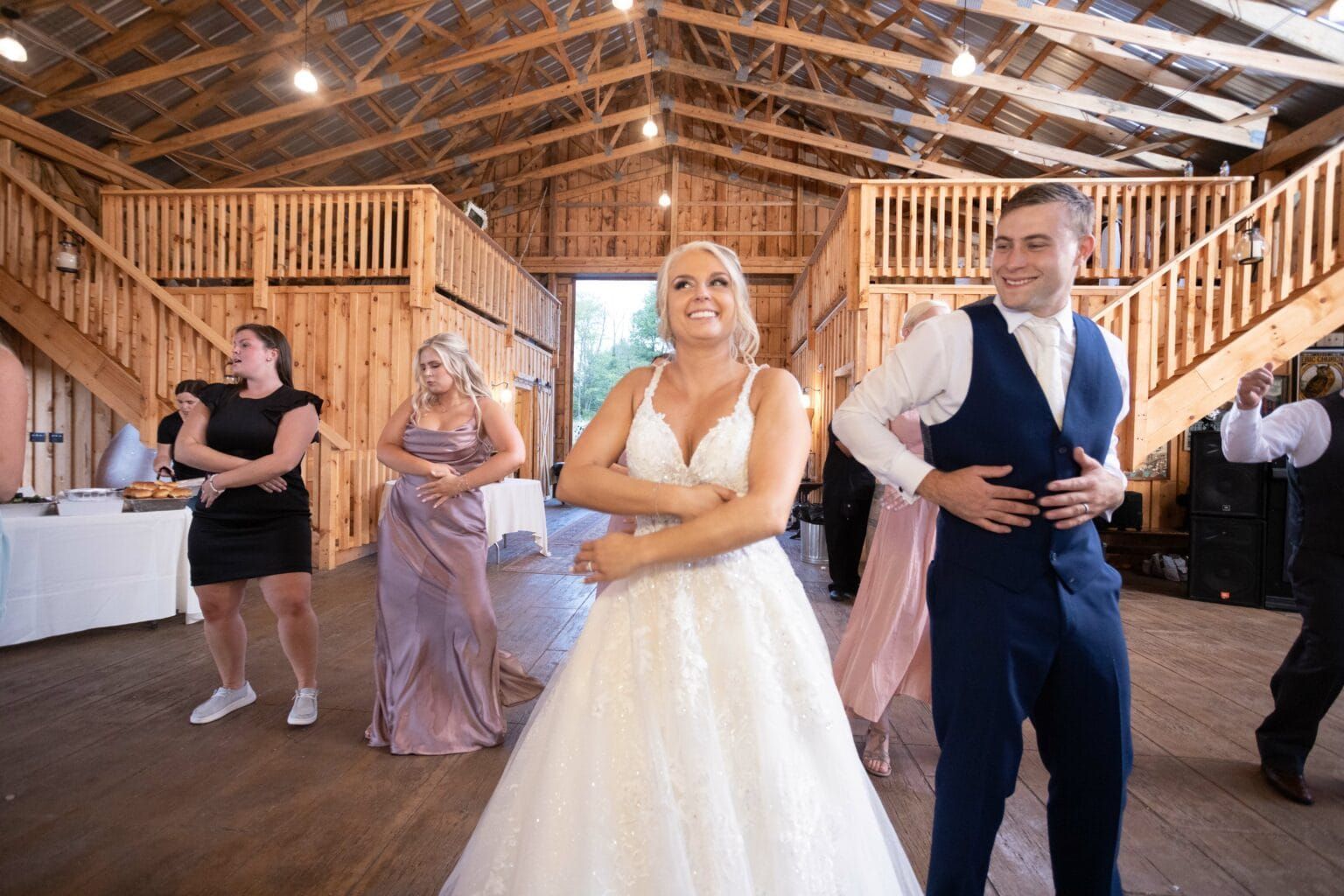 A bride and groom are dancing with their wedding party in a barn.