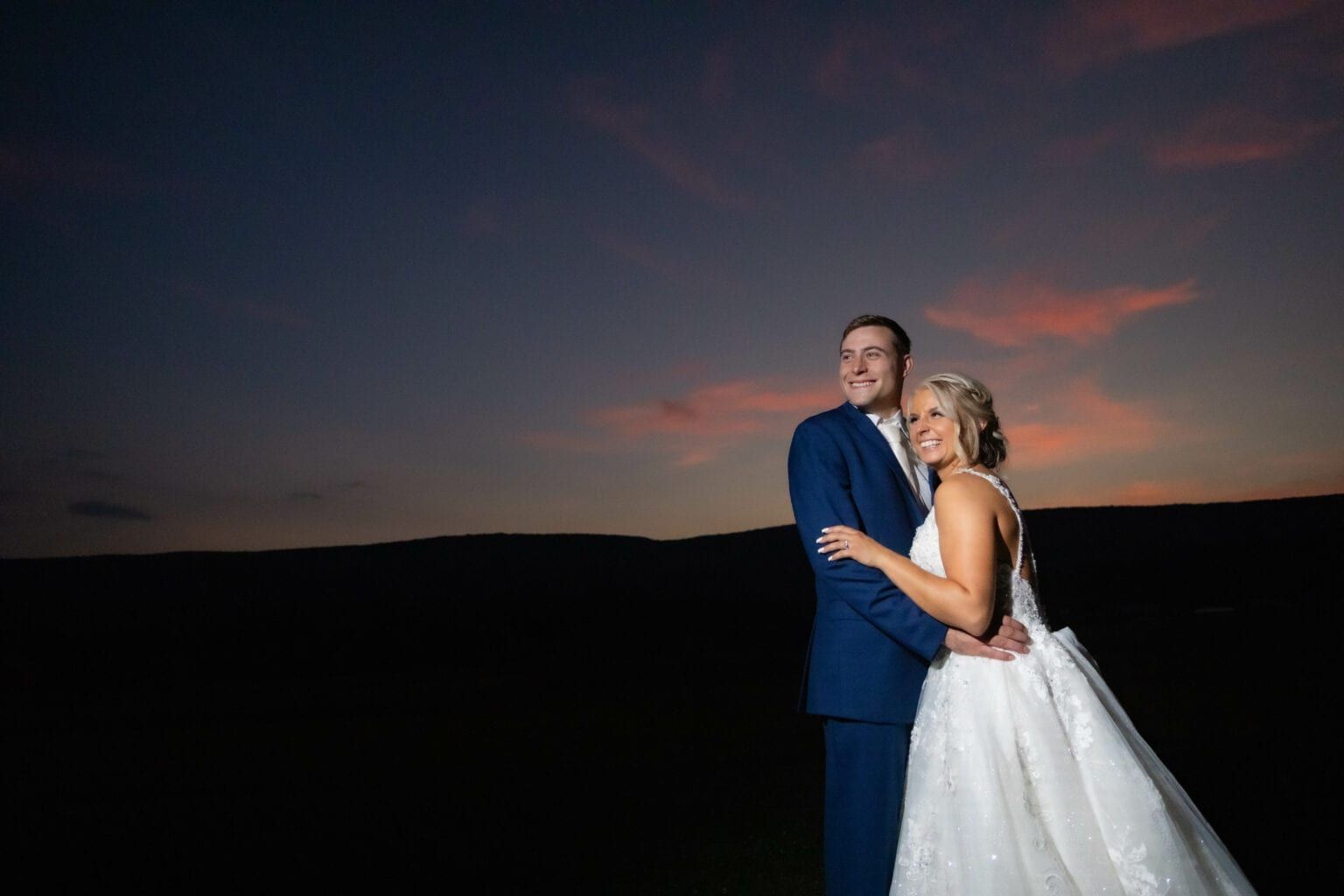 A bride and groom are standing next to each other in a field at sunset.