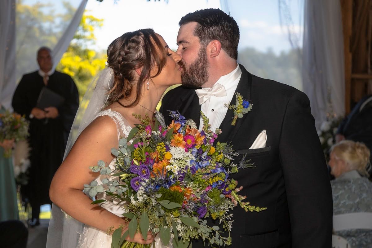 A bride and groom kissing at their wedding ceremony while holding a bouquet of flowers.