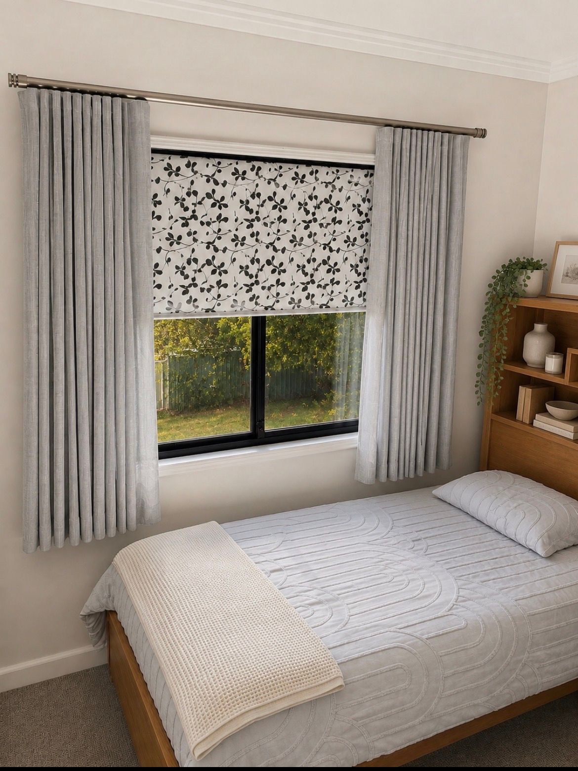 Bright bedroom with a bed, window and gray curtains, patterned blind, and wooden shelf with plants— Curtain Concepts in Dunbible, NSW