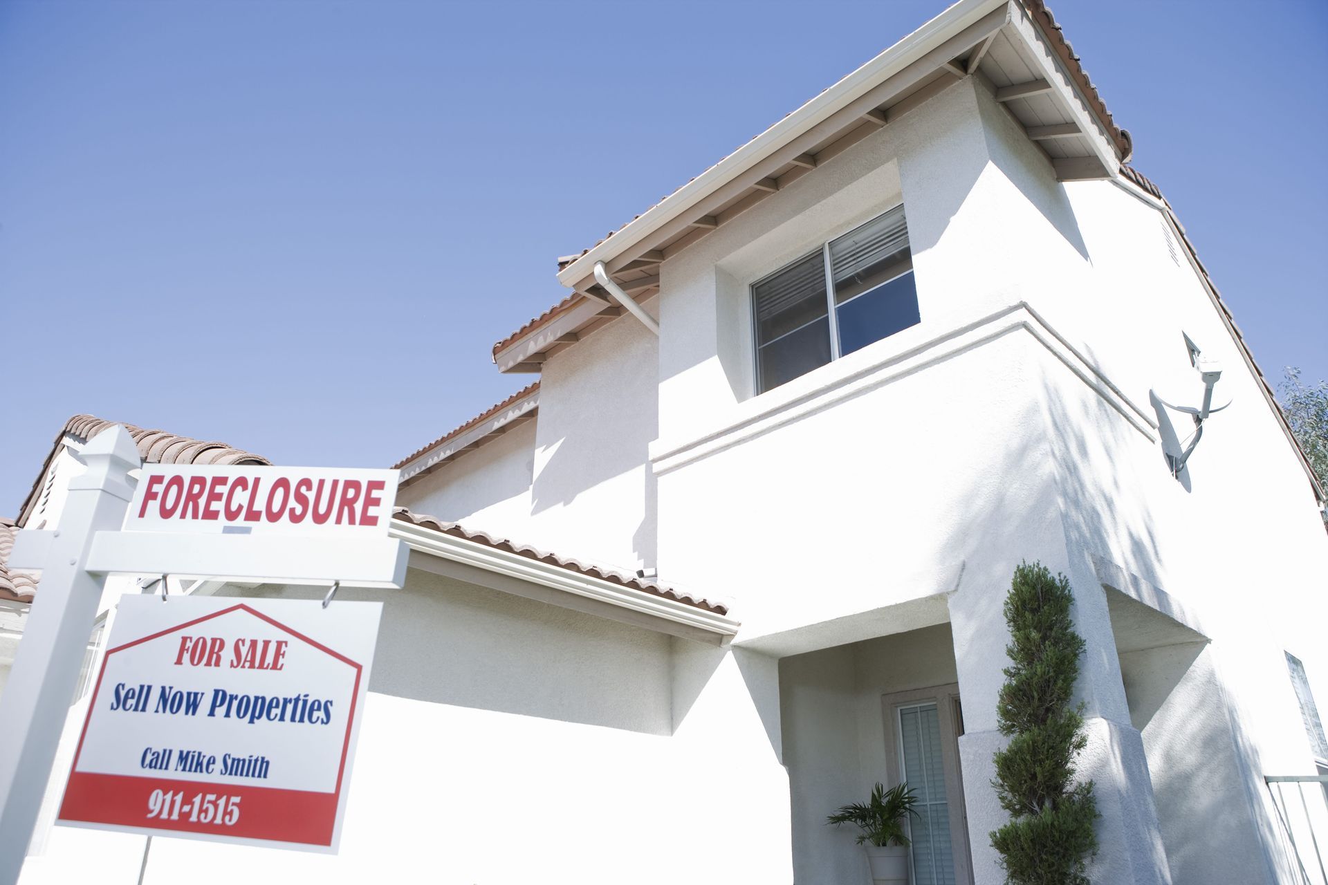Foreclosure sign in front of a white two-story house under a clear blue sky