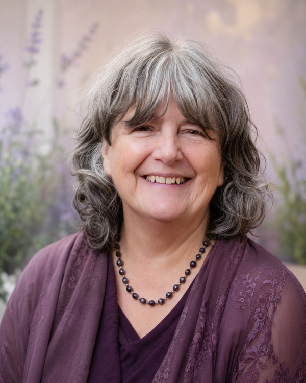 Smiling person with gray wavy hair wearing a purple top and beaded necklace against a soft floral background