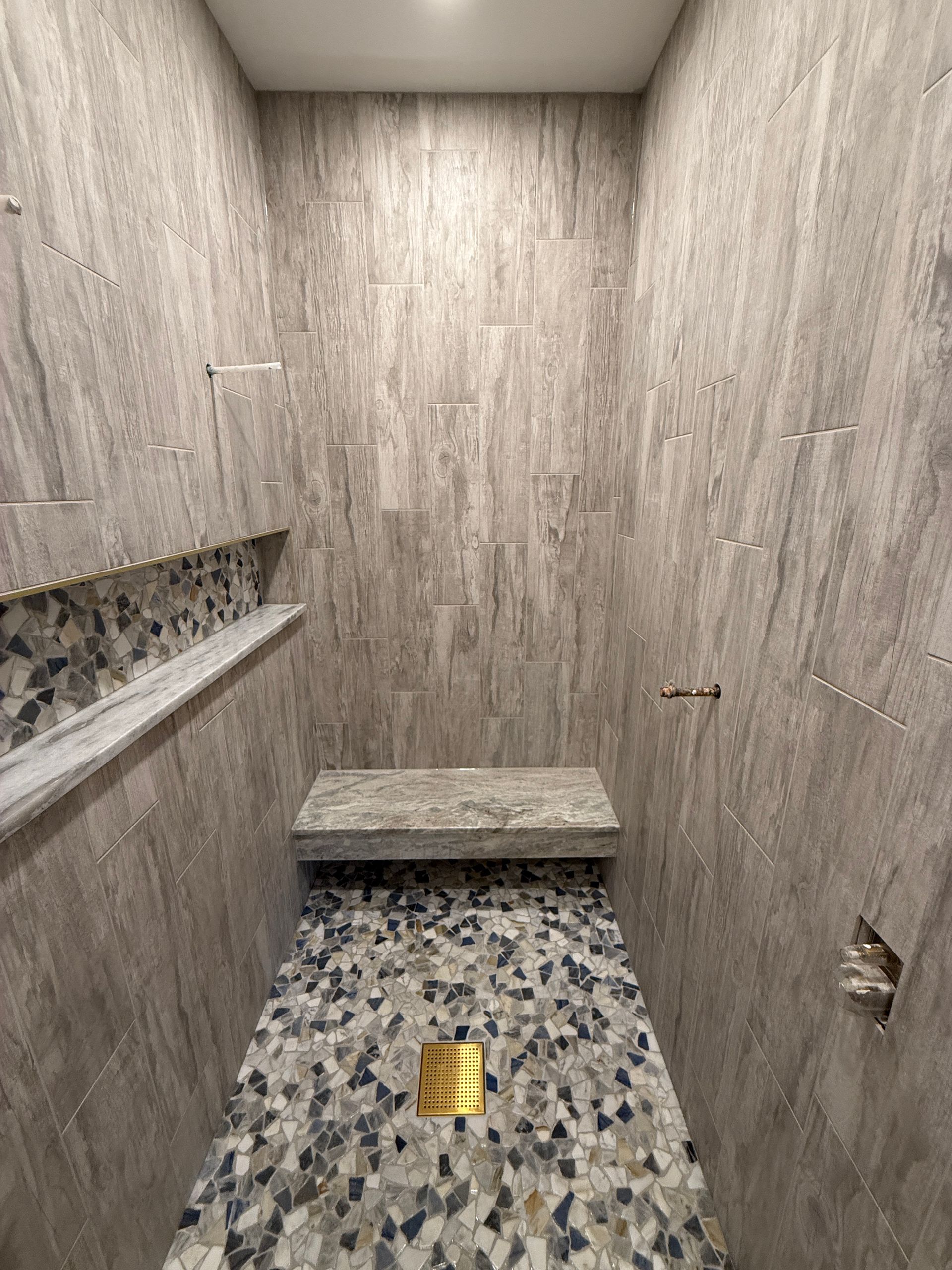 Shower with gray tiled walls, stone bench, and pebble mosaic floor with a gold drain.