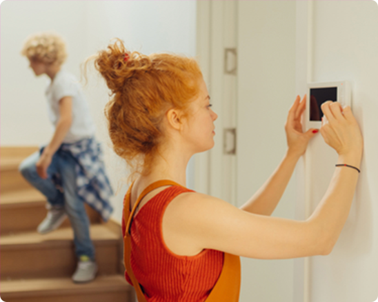 A woman is adjusting a thermostat on a wall while a boy sits on the stairs.