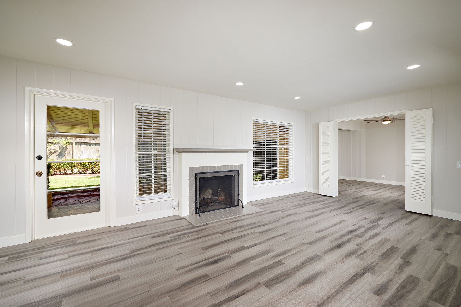 Bright empty living room with gray wood floors, white walls, fireplace, and shuttered windows